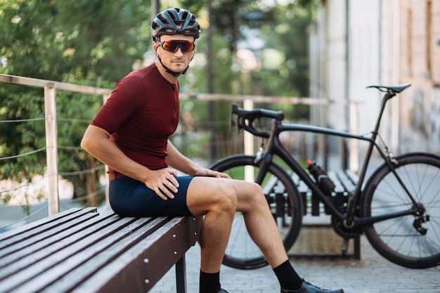 黒い自転車の近くの木製のベンチに座っているスポーツウェアのヘルメットと眼鏡の筋肉サイクリスト屋外で休んでいる間カメラを見ている白人男性