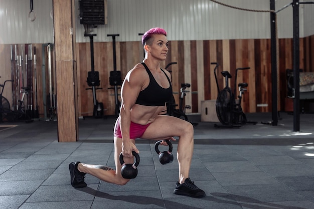 Muscular extraordinary female athlete with short pink hair trains lunges with heavy kettlebells. Functional, cross training in modern gym.