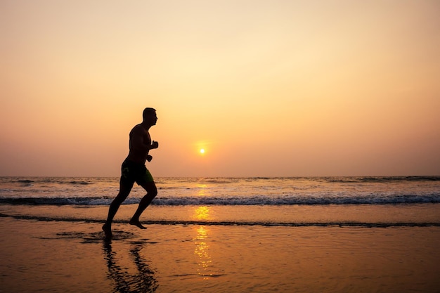 운동을 하는 근육질의 건강한 젊은 보디빌더 남자는 일몰 시 파라다이스 해변에서 스트레칭을 하고 있습니다. 바다에서 피트니스 남성 모델 스포츠