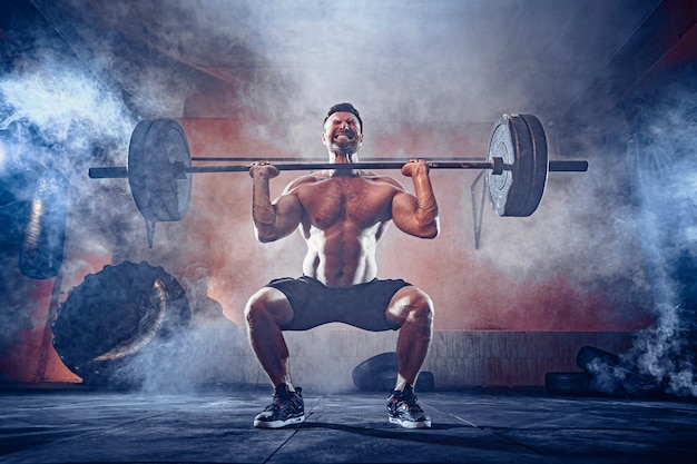 Muscular fitness man doing deadlift a barbell over his head in modern fitness center. Functional training. Snatch exercise. Smoke on wall.