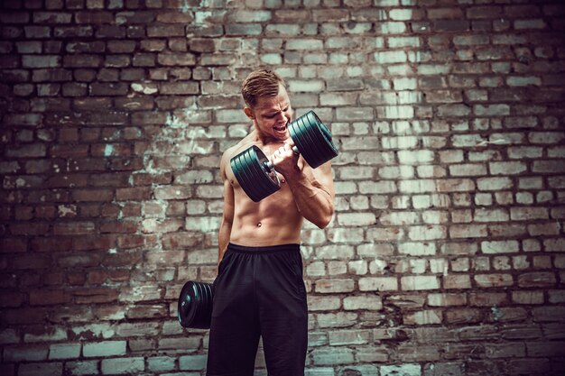 Muscular guy doing exercises with dumbbell against a brick wall