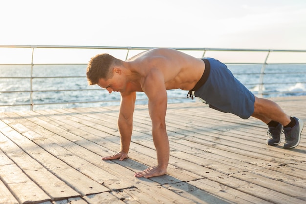Muscular handsome fitness man with naked torso doing push-ups on the beach at sunrise