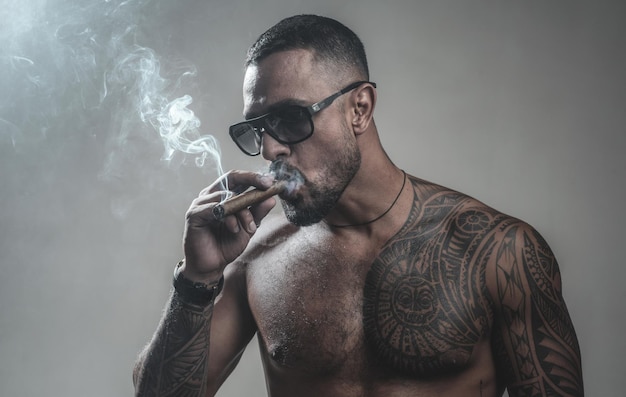 Muscular macho man with athletic body sexy abs of tattoo man sport and fitness health confidence charisma brutal sportsman torso steroids smoking cigarette exhale smoke bad habit harmful