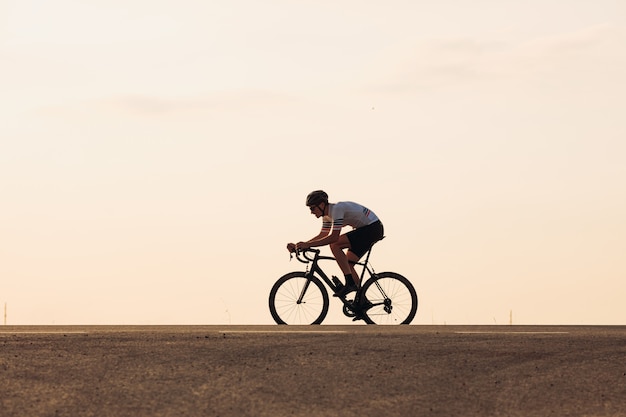 Muscular male athlete in activewear and protective helmet cycling on asphalt road during sunset