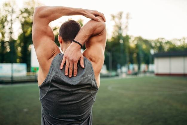 Muscular male athlete doing stretching exercise, back view, fitness workout. Strong sportsman in park