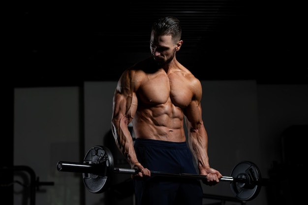 Photo muscular man doing heavy weight exercise for biceps with barbell in gym