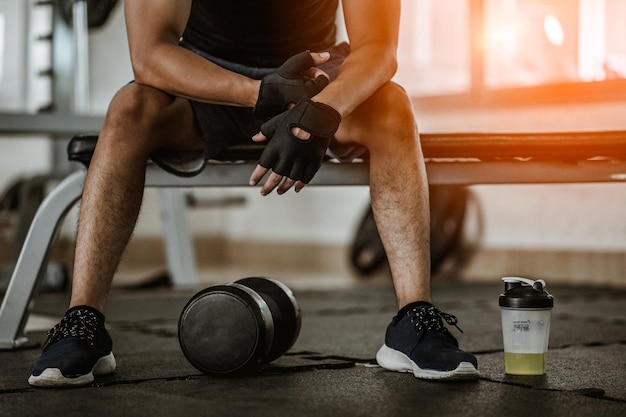 Muscular man at gym taking a break from workout sitting beside the bottle of mineral salt and dumbbells .