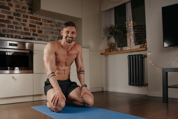 A muscular man is sitting on a blue yoga mat in his apartment