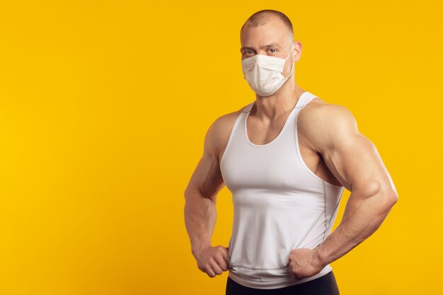 Muscular man in a medical mask and white shirt, standing over isolated yellow wall