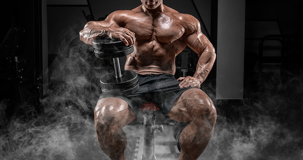 Muscular man sits on a bench in smoke with dumbbells. Bodybuilding and powerlifting concept. Mixed media