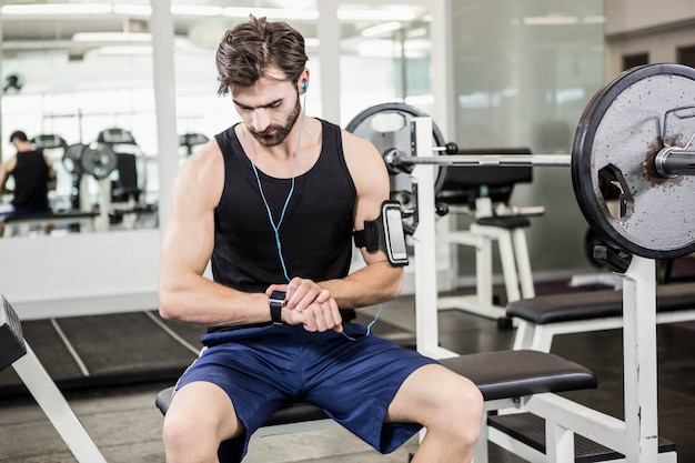 Uomo muscolare che si siede sul banco del bilanciere e utilizzando smartwatch in palestra