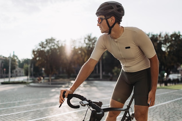 Muscular man in sport clothes helmet and glasses sitting on bike outdoors and looking aside Caucasian sportsman taking break after cycling on city street