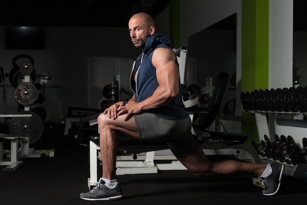 Muscular man stretching his leg at the floor in a gym and flexing muscles  muscular athletic bodybuilder fitness model