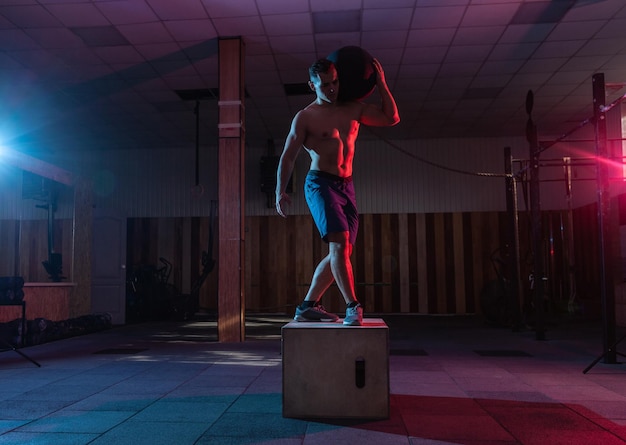 Muscular man training wooden box pacing with a heavy bag in dark gym Athlete workout in red blue neon light Healthy lifestyle cross training