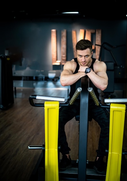 Muscular man trains in the gym man sits on simulator and looks at the camera