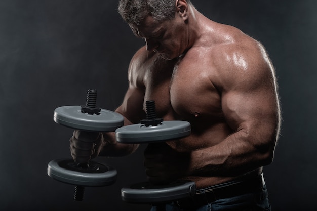 Muscular man trains with dumbbells on black background in smoke