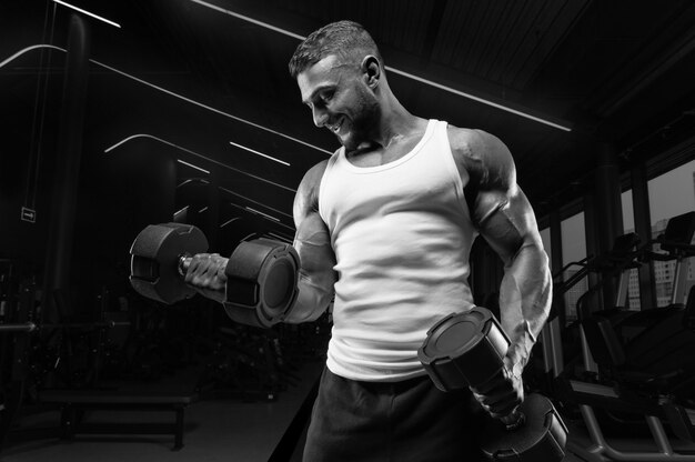 Muscular man in a white t-shirt works out in the gym with dumbbells. Biceps pumping. Fitness and bodybuilding concept.