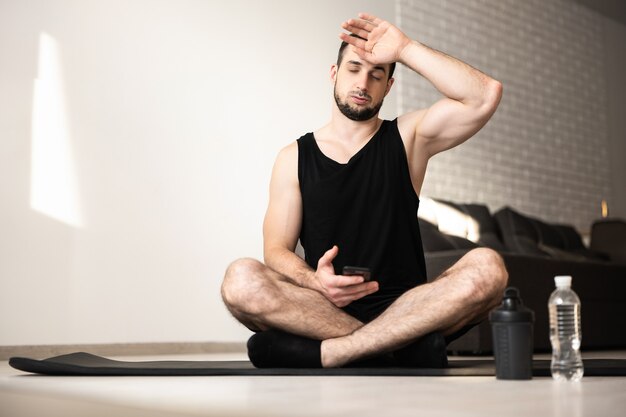 Muscular man wipes sweat of his forehead after hard home workout. Healthy lifestyle concept. Man tired after intensive training. Plastic water bottle. Light living room on background.