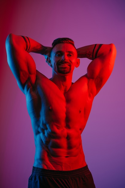 A muscular man with a beard is resting with arms behind his head under the blue and red light