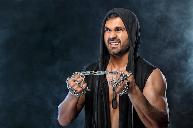 Muscular man with chain on black background with smoke. Strong bodybuilder try to break chain