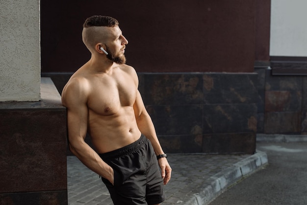 Muscular man with naked torso wearing an earphones is posing against the concrete wall outdoors