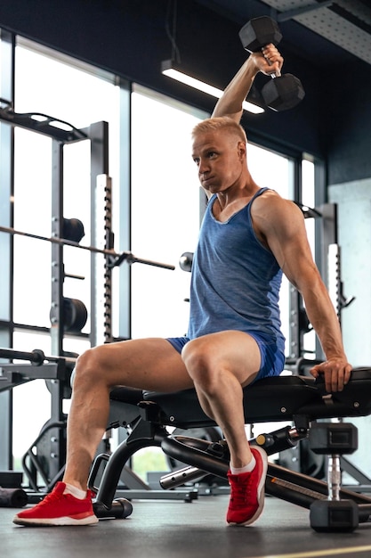 Мускулистый мужчина тренируется в тренажерном зале, делает упражнения с гантелями на трицепс