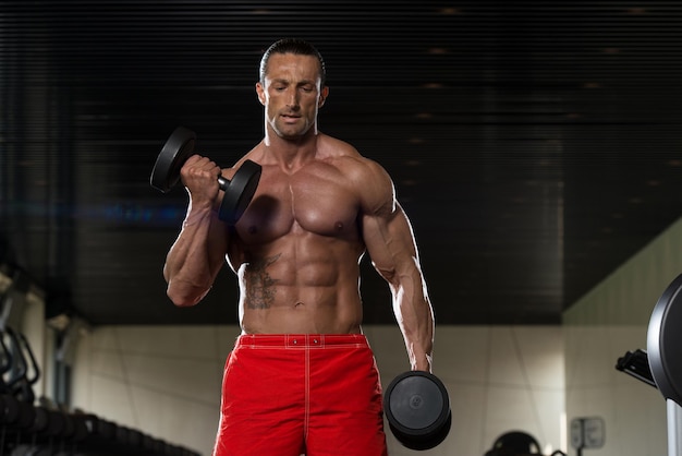 Мускулистый взрослый мужчина делает тяжелые упражнения для бицепсов с гантелями в современном фитнес-центре