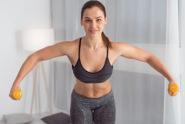 Muscular. Pretty joyful well-built dark-haired young woman smiling and holding hand weights while doing exercises for arms at home