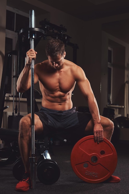 A muscular shirtless athlete holds barbell disc while sits on a bench in the gym.