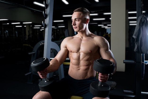 A muscular shirtless man is holding dumbbells on his knees in a gym.