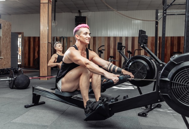 Muscular sporty women workout on training simulator in crossfit gym. Sportswomen exercising on rowing machines