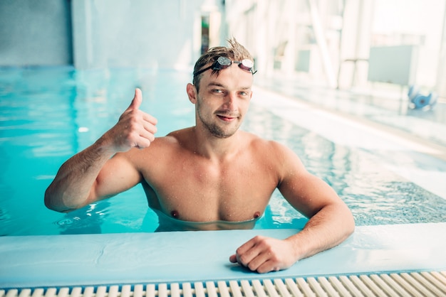 Muscular swimmer shows thumbs up in indoor swimming pool.