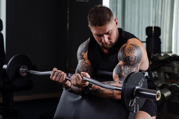 Muscular tattooed athlete swings his biceps with a barbell in the gym