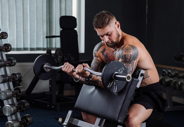 Muscular tattooed athlete swings his biceps with a barbell in the gym