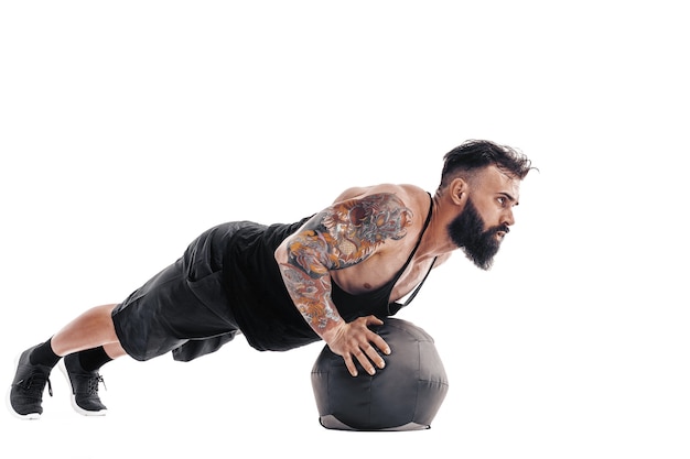 Muscular tattooed bearded male exercising fitness weights Medicine Ball push ups exercises in  isolated on white wall.