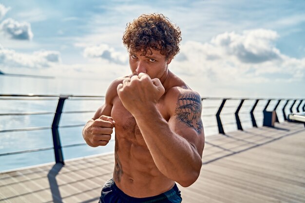 Muscular young man athlete standing and practicing shadow boxing outdoors early in the morning on pi...