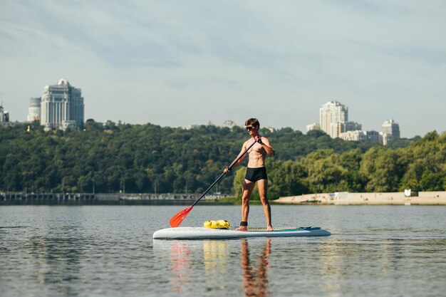 Muscular young man stands on sup board on water and paddles on background of beautiful landscape