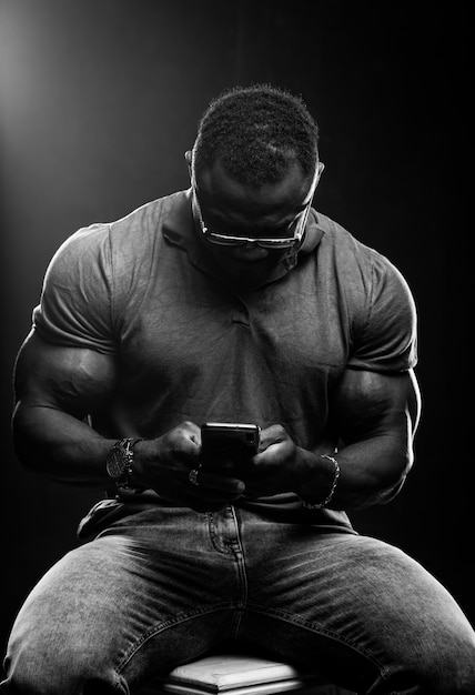 Muscular young man types on phone with head down Unrecognizible strong male on high bar chair Black and white portrait