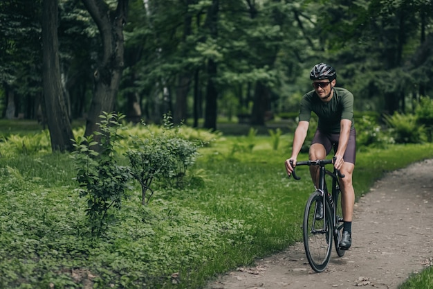녹색 도시 공원에서 운동복, 보호 헬멧, 거울 안경을 쓴 근육질의 청년이 자전거를 타고 있습니다. 백인 남자의 야외 활동입니다. 건강한 라이프 스타일.