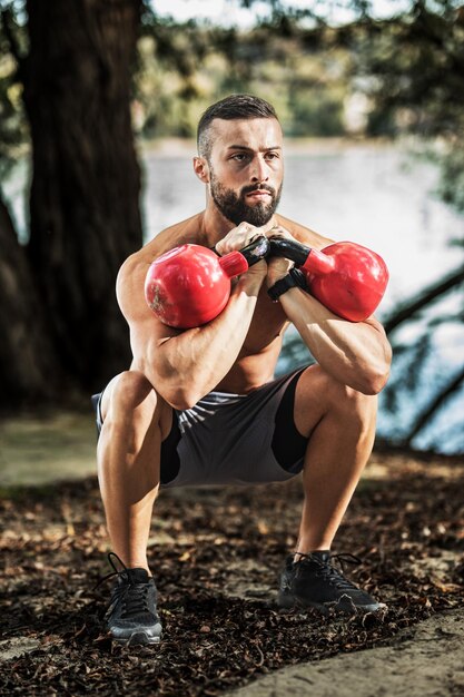 Giovane muscolare con il torso nudo che fa esercizi di squat con kettlebell nella natura, vicino al fiume.