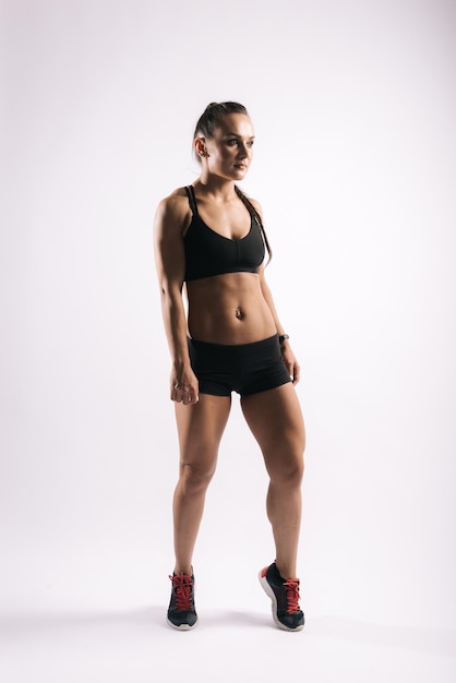 Muscular young woman with perfect athletic body in sports black bra is standing and posing on white isolated background