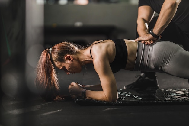Мускулистая молодая женщина тренируется с личным тренером в тренажерном зале. Она делает упражнения на доске.
