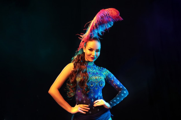 Music. Beautiful young woman in carnival, stylish masquerade costume with feathers on black background in neon light. Copyspace for ad. Holidays celebration, dancing, fashion. Festive time, party.