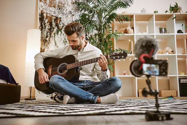 Photo music blogger sitting on the floor and holding the guitar, while recording new video for his vlog.