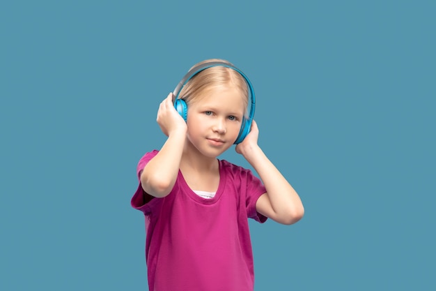 Music, headphones. Cute blonde girl in bright tshirt in blue headphones in great mood in photo studio