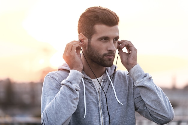 Music is always with me. Confident young man putting headphones into his ears and looking away while standing outdoors