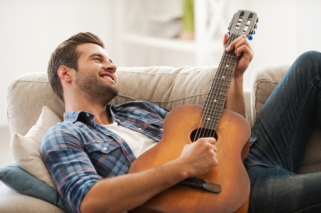 音楽は私の人生です。ソファに横になってギターを弾く幸せな若い男
