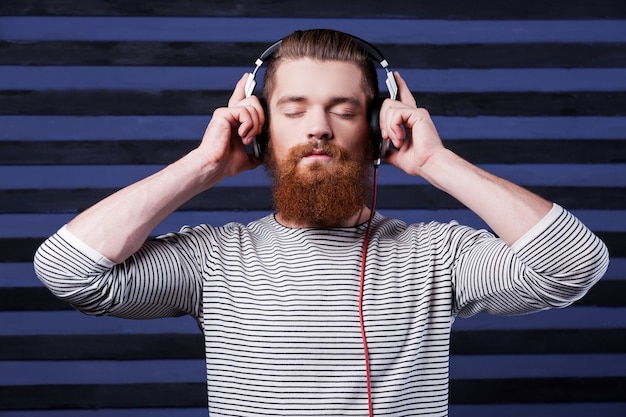 Music lover. Confident young bearded man in headphones standing against stripped background and listening to the music