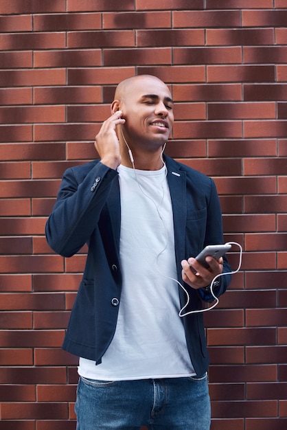 スマートフォンで音楽を聴いて壁に立っているイヤホンを身に着けている音楽愛好家の若い男は閉じました