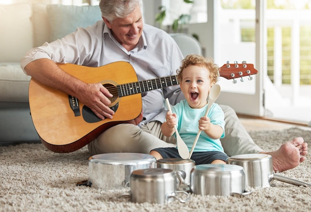 ミュージック ポットとベビー ドラマーの老人とリビング ルームの床に鍋と木のスプーンの楽器と彼のギター 思い出の笑顔と年配の祖父母は幸せな孫との時間を楽しんでいます